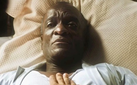 O ator Christovam Neto está deitado em uma cama e tem uma mão no seu peito em cena da novela Amor Perfeito