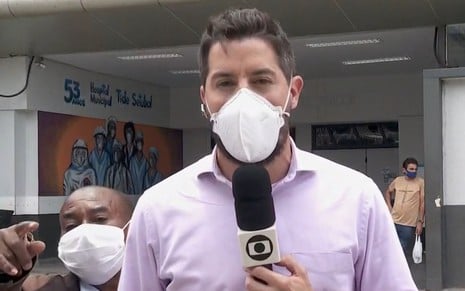 Repórter Anselmo Caparica em frente ao hospital Tíde Stúbal. Ele está de máscara, com o microfone na mão direita e veste uma camisa branca. Ao seu lado direito é possível ver o homem que o interrompeu durante a transmissão.