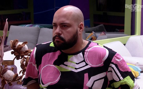 Tiago Abravanel com cara séria dentro da casa do BBB; ele veste camisa preta com estampas grandes rosa e branco