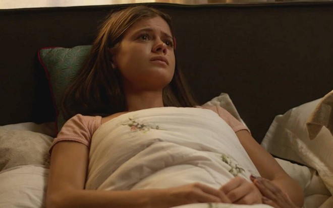 Sara Vidal surge deitada em cama, junta as mãos e olha para cima em cena de Quanto Mais Vida, Melhor