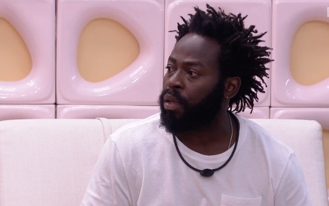 O ator Douglas Silva, com camisa branca, sentado em um sofá branco na área externa do Big Brother Brasil 22