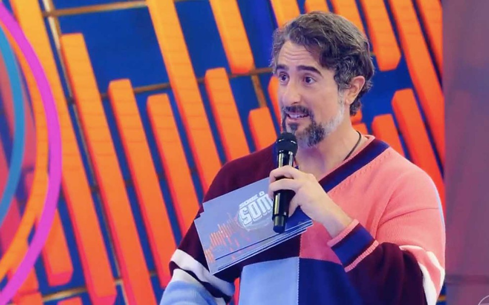 Marcos Mion segura uma ficha com o logo do Sobe o Som e um microfone preto com a mão direita no palco do Caldeirão