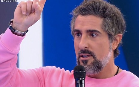 O apresentador Marcos Mion, de camiseta rosa, ergue o dedo indicador da mão esquerda para o alto, no palco do Caldeirão
