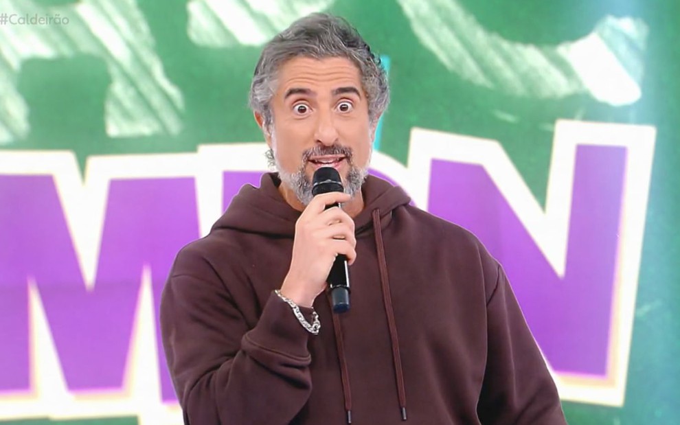 O apresentador Marcos Mion segura um microfone com a mão direita enquanto está com a expressão com olhos arregalados