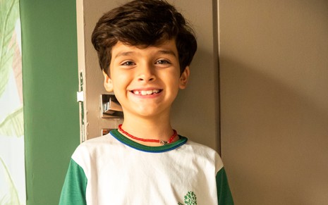 Guilherme Tavares sorri para a câmera em ensaio de divulgação de Cara e Coragem; ele veste um uniforme escolar branco, com detalhes em verde. Na camiseta, é possível observar a inscrição "Escola Jardim".