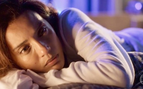A atriz Taís Araujo está deitada e apoia a cabeça em um dos braços em cena da novela Cara e Coragem; ela usa uma pijama e tem olhar triste