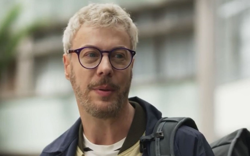 Jonathan (Guilherme Weber) está em pé, usa óculos e está sério em cena de Cara e Coragem, novela das sete da Globo