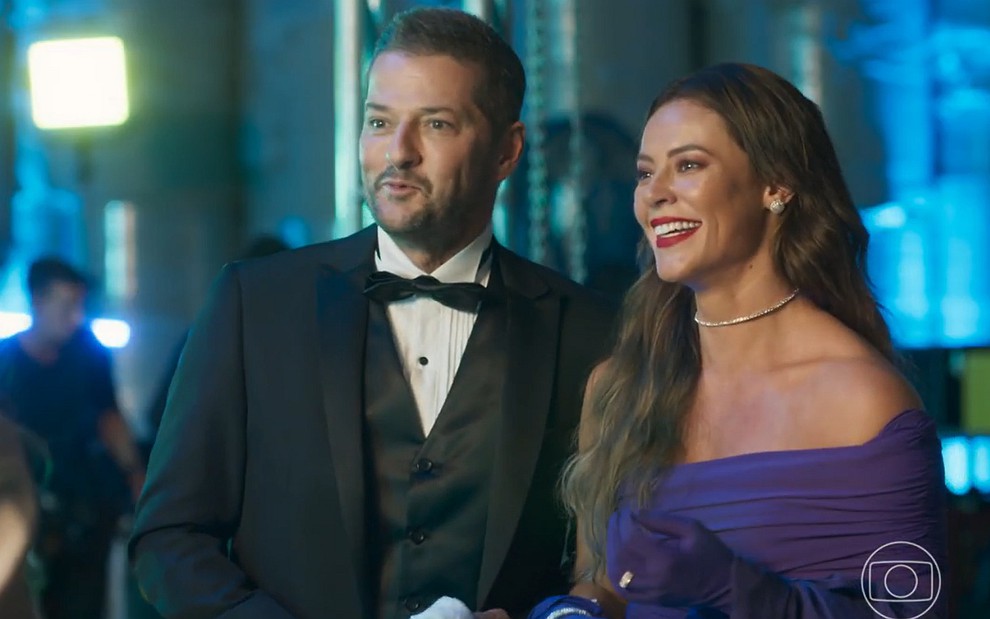 Marcelo Serrado e Paolla Oliveira sorriem vestidos de roupas de gala na novela Cara e Coragem