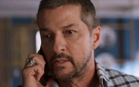 O ator Marcelo Serrado está em close com um celular na orelha em cena da novela Cara e Coragem