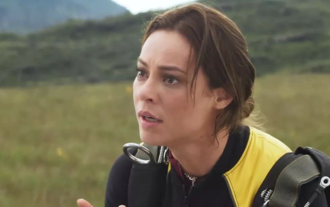 Paolla Oliveira veste uma roupa específica de paraquedismo e usa um coque frouxo; a atriz tem a expressão perturbada em cena de Cara e Coragem