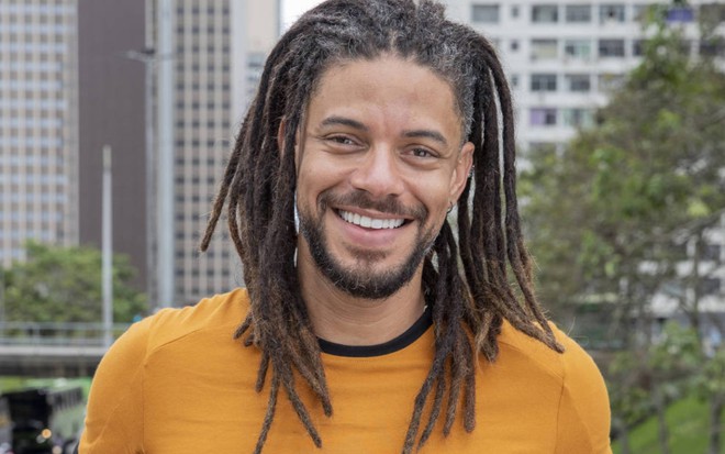 Paulo Lessa usa longos dreadlocks e barba; ele sorri para a câmera em ensaio fotográfico de Cara e Coragem