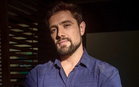 O ator Rafael Cardoso está em um local escuro e usa uma camisa azul marinho caracterizado como Rômulo da novela Cara e Coragem