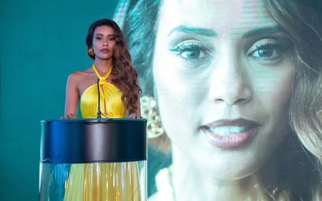 Taís Araujo usa um vestido decotado, amarelo, e posa atrás de um palanque; ela tem a expressão séria. Ao fundo, é possível ver uma imagem dela projetada.