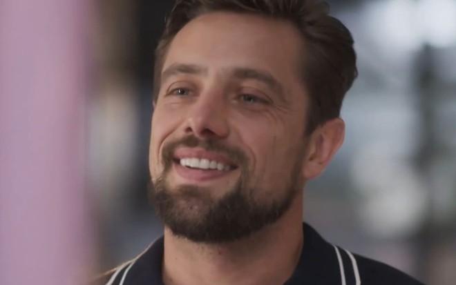 Rafael Cardoso usa uma camisa polo preta com listras brancas; ele dá um sorriso sedutor em cena de Cara e Coragem
