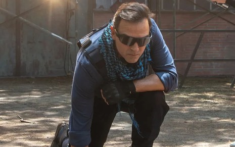 Tadeu Schmidt está agachado, com botas, camisa azul, cachecol e óculos escuros, e tem o semblante concentrado em cena de Cara e Coragem