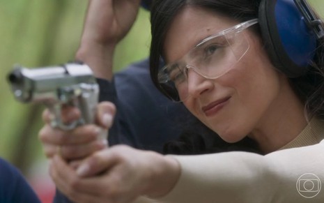 Mel Lisboa está com óculos de proteção e aponta revolver como se fosse atirar caracterizada como Regina em Cara e Coragem