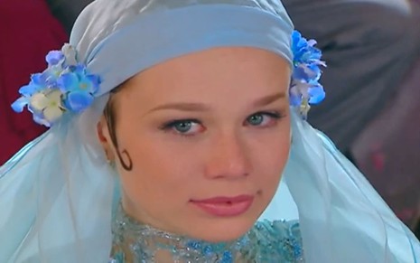 Mariana Ximenes usa um vestido de noiva típico da década de 1920, com rendas e flores artificiais; o traje inteiro, inclusive o véu, é azul celeste