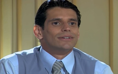 Alexandre Barillari usa camisa social colete e gravata cinzas e tem os cabelos cheios de gel; ele dá um leve sorriso em cena de Chocolate com Pimenta