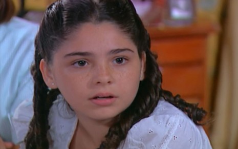 Aos 9 anos, Marcela Barrozo tem os cabelos cacheados e usa um vestido rodado amarelo; ela tem o semblante sério em cena de Chocolate com Pimenta