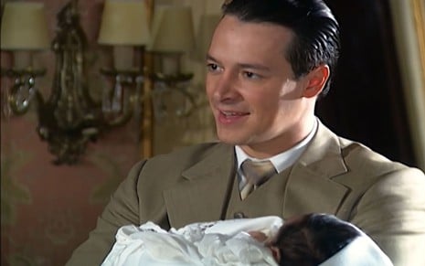 Rodrigo Faro caracterizado como Guilherme; ele usa um terno cinza, sorri e segura um bebê em cena de Chocolate com Pimenta