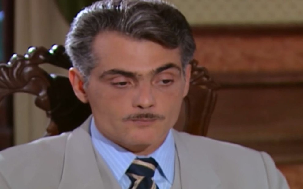 Tarcisio Filho, caracterizado como Sebastian, dá um sorriso irônico em cena de Chocolate com Pimenta; ator tem cabelos e bigode grisalhos e usa um terno cinza