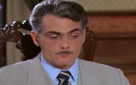 Tarcisio Filho, caracterizado como Sebastian, dá um sorriso irônico em cena de Chocolate com Pimenta; ator tem cabelos e bigode grisalhos e usa um terno cinza