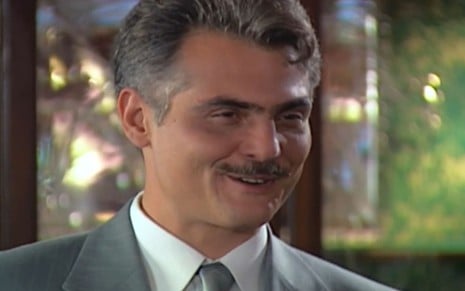 Tarcísio Filho, caracterizado como Sebastian, dá um sorriso irônico em cena de Chocolate com Pimenta; ator tem cabelos e bigode grisalhos e usa um terno cinza