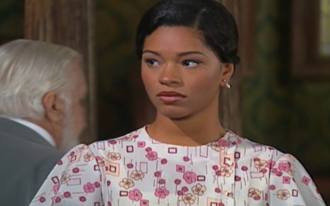 Juliana Alves, caracterizada como Selma, tem a expressão abalada e tristonha; ao fundo, é possível ver Cláudio Corrêa e Castro de passagem