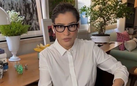 Renata Vasconcellos usa uma camisa social branca, um óculos de aro preto e cabelos amarrados. Ela encara a câmera, sorridente, durante o Conversa com Bial