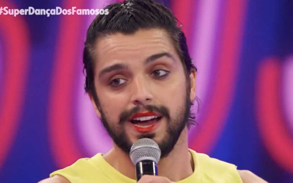 O ator Rodrigo Simas com um cropped amarelo, ele usa um batom vermelho, no palco da Super Dança dos Famosos