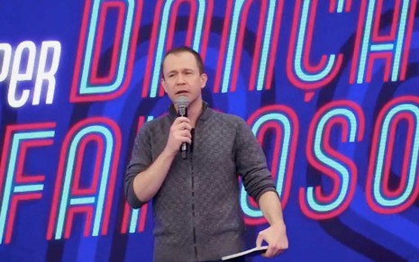 O apresentador TIago Leifert com um microfone na mão direita e cenho franzido em frente a um telão em que se lê Super Dança dos Famosos