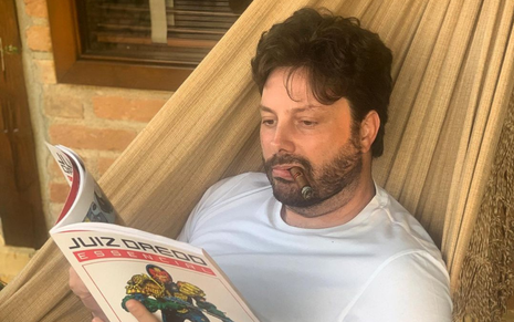 Danilo Gentili deitado na rede, lendo um livro e fumando um charuto