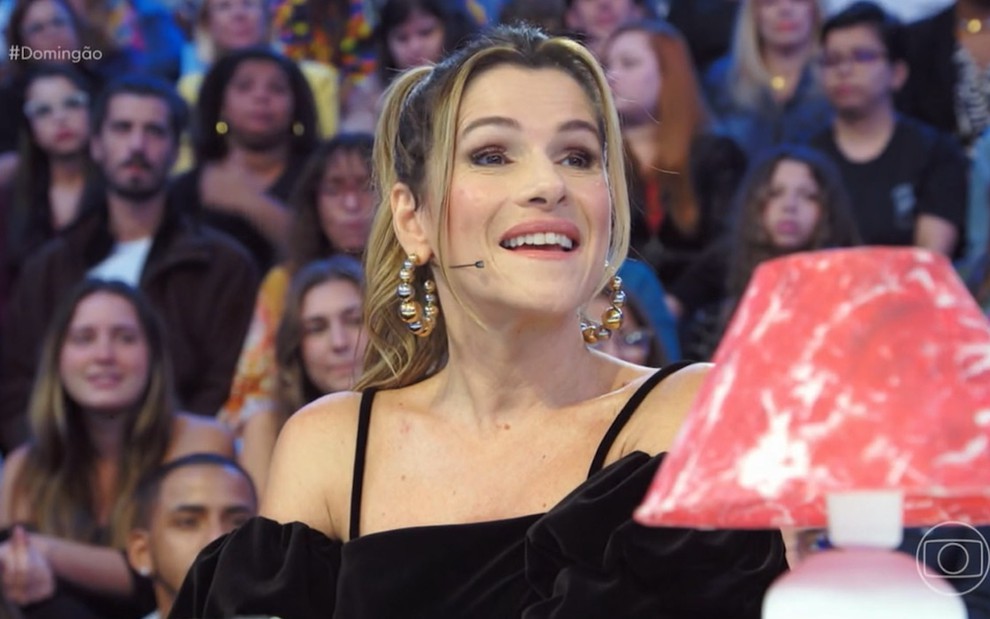 A humorista Ingrid Guimarães está com a boca entreaberta olhando para o lado, com plateia do Domingão com Huck, ao fundo