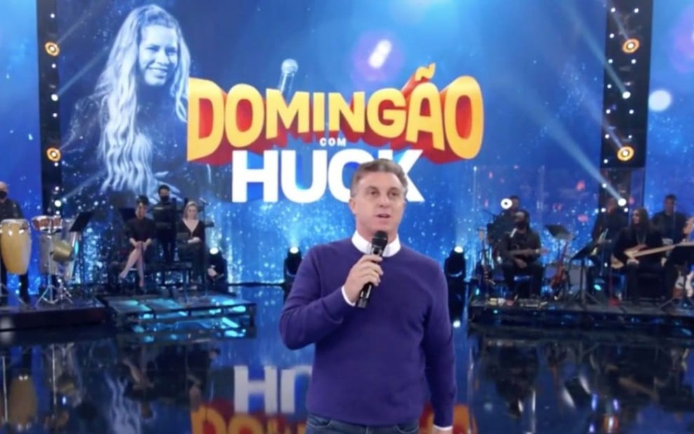 O apresentador Luciano Huck no Domingão com Huck de domingo (7), com imagem de Marília Mendonça no telão do programa