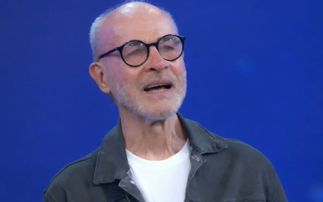 Osmar Prado está com a cabeça raspada e a barba rala; ele dá um leve sorriso enquanto encara o público durante participação no Domingão com Huck