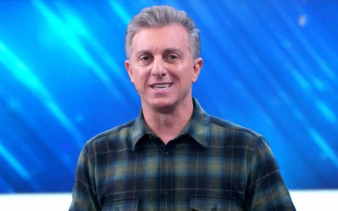 Luciano Huck sorri forçadamente no cenário do Domingão com Huck