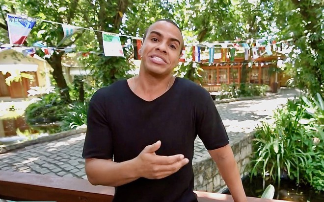 O jornalista Thiago Oliveira usa camisa preta e está em um jardim do cenário do programa É de Casa