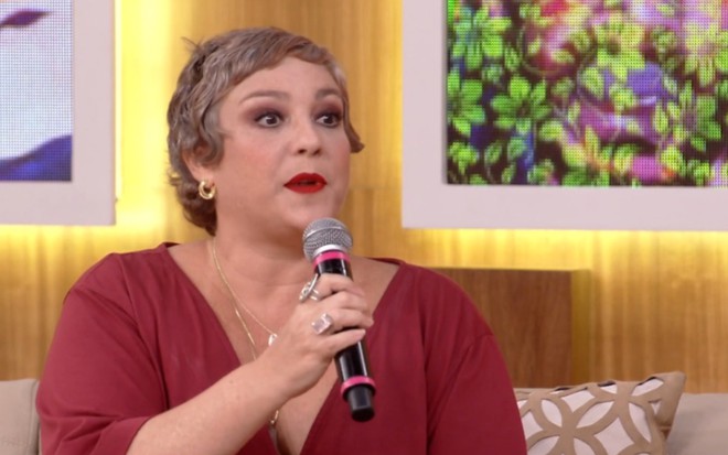 Ana Baird usa cabelos curtos, grisalhos, e um macacão vermelho terroso. Ela segura um microfone em frente ao cenário do Encontro e responde as perguntas de Patrícia Poeta