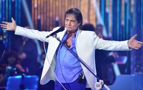 Roberto Carlos abre os braços no palco, ele veste blazer branco e camisa azul