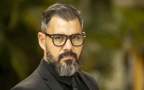Juliano Cazarré está com terno, camisa e gravata escuros, usa óculos e tem barba e cabelos bem penteados como Pascoal da novela Fuzuê