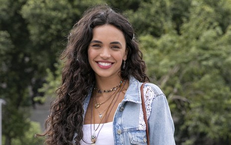 A atriz Giovana Cordeiro usa jaqueta jeans, top branco, acessórios e sorri caracterizada como Luna, mocinha de Fuzuê