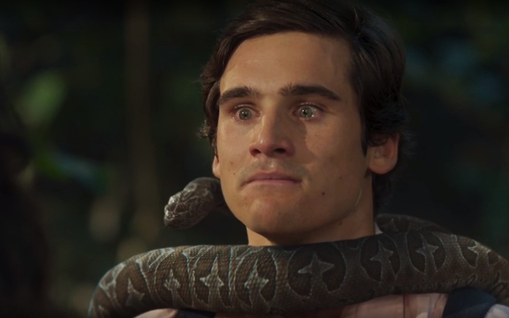 O ator Nicolas Prattes está paralisado com uma cobra enrolada no pescoço em cena da novela Fuzuê como Miguel