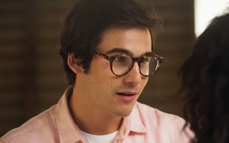 Nicolas Prattes caracterizado como Miguel; ele usa um óculos de aro preto e cabelo curto e bem alinhado. O semblante exprime susto em cena de Fuzuê.