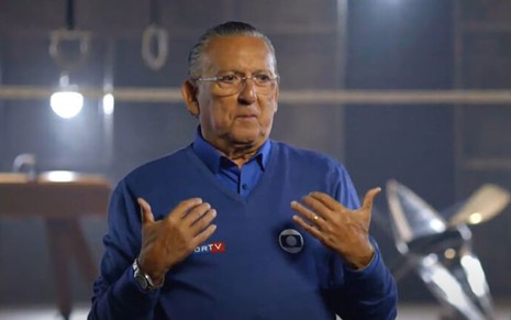 Galvão Bueno na chamada para os Jogos Olímpicos, com uma camisa azul, apontando para si mesmo, em um fundo com palcos de esportes olímpicos