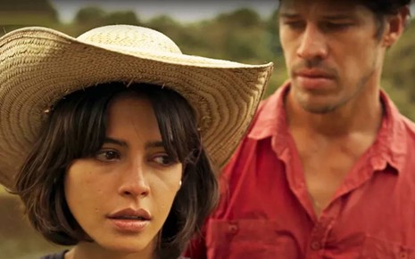 Atriz Julia Dalavia olha para o lado com cara de séria, enquanto José Loreto aparece ao fundo, em cena de Pantanal