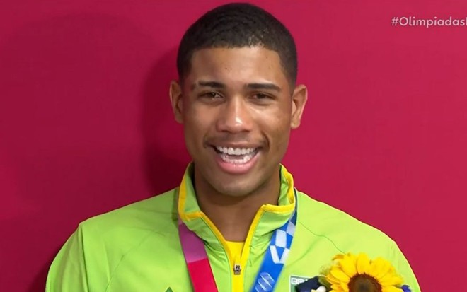 Hebert Conceição em entrevista para Globo: atleta sorria, usa blusão verde limão e está com o cordão da medalha ao redor do pescoço