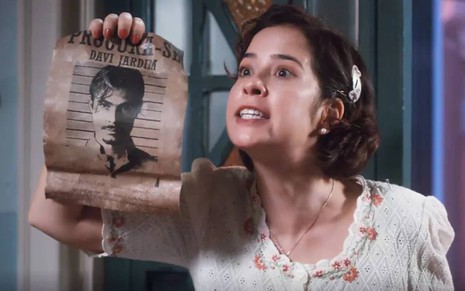 Atriz Paloma Duarte segura cartaz com o rosto de Rafael Vitti e olha para frente com cara de nervosa em cena de Além da Ilusão