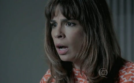 Maria Ribeiro grava com camisa vermelha e branca, cabelo solto com franja e expressão de choque como Danielle de Império