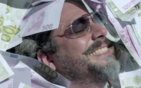 O ator Alexandre Nero grava cena de Império sorrindo, com óculos escuros, com apenas parte do rosto aparecendo em meio à notas de euros