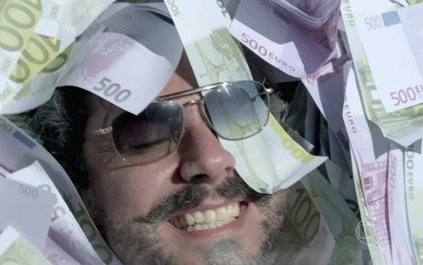 Alexandre Nero grava com óculos de sol, sorrindo, e com notas de dinheiro na cara como José Alfredo em Império